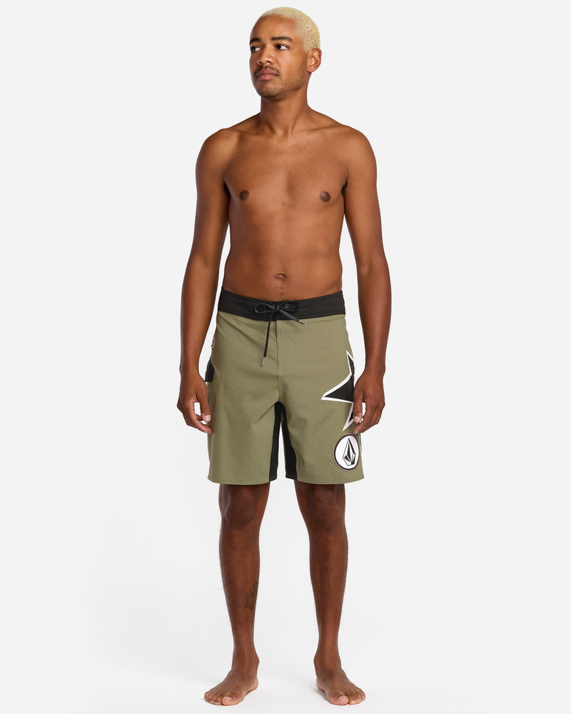 A shirtless, barefoot man with light blonde hair models Volcom Men's St4R G4Zer Mod 20 Trunks in olive green and black with a white geometric design, set against a plain white background.