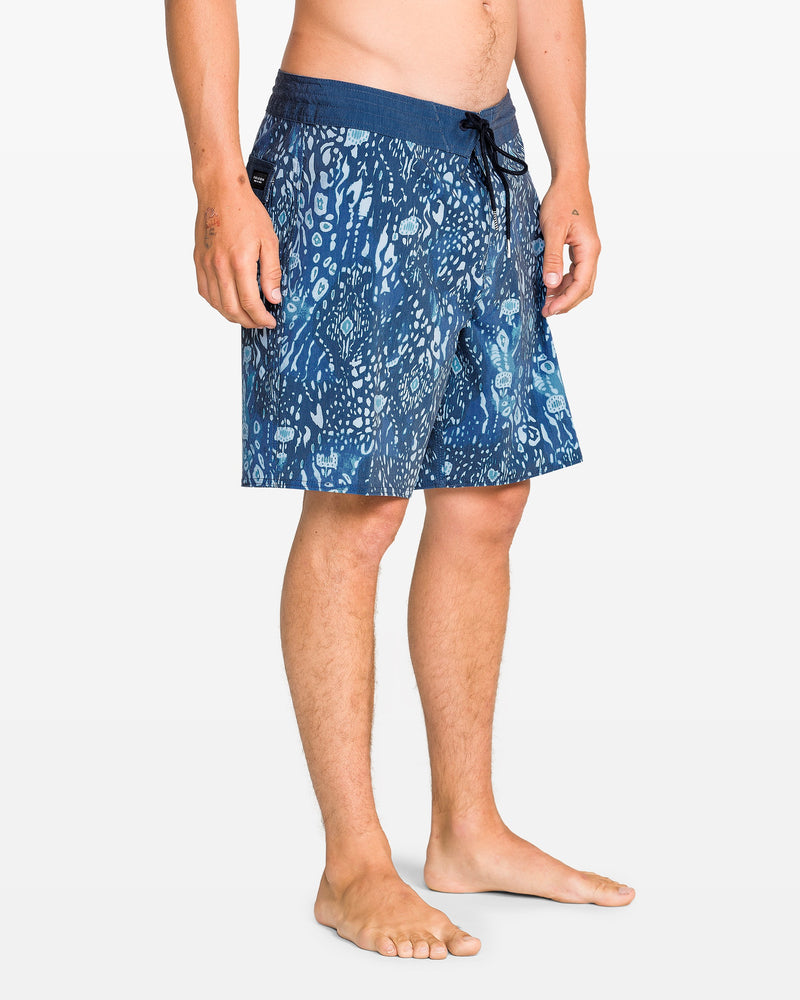 A man stands barefoot in Volcom Men's Psyche Cotton 17" Trunk, featuring blue patterns and a drawstring waist, shown from mid-torso to feet against a plain white background.
