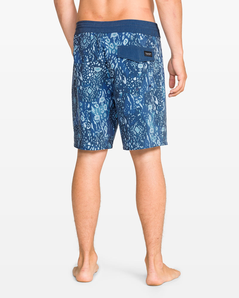 A man stands barefoot, facing away, wearing Volcom Men's Psyche Cotton 17" Trunk boardshorts with blue patterns, a dark waistband, and a right-side back pocket.