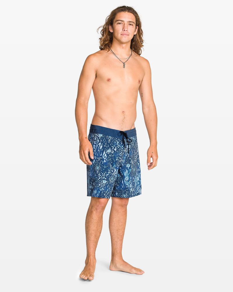 A young man with long hair and a relaxed expression stands barefoot on a white background, wearing Volcom men's Psyche Cotton 17" Trunk boardshorts and a necklace. He is shirtless and faces slightly to the side.