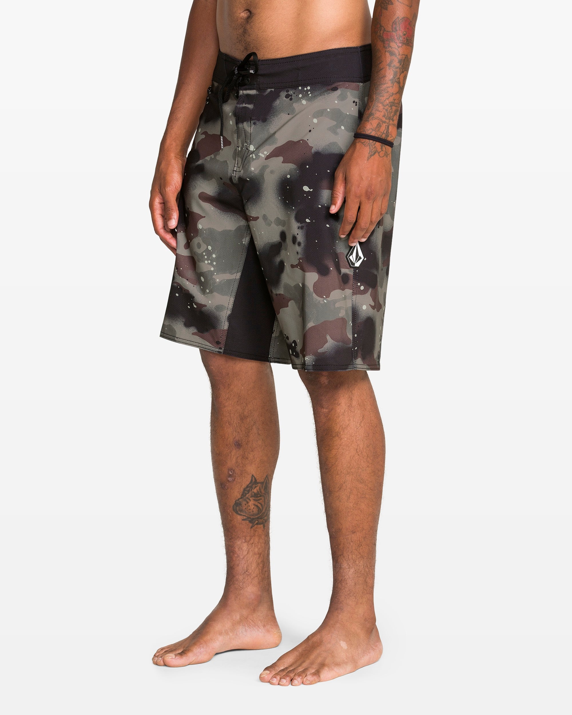 A person stands barefoot in Volcom Men's Fomenter Mod black and gray camo board shorts with a drawstring waist, made from REPREVE recycled fibers. Their left arm and leg have tattoos. The background is plain white.