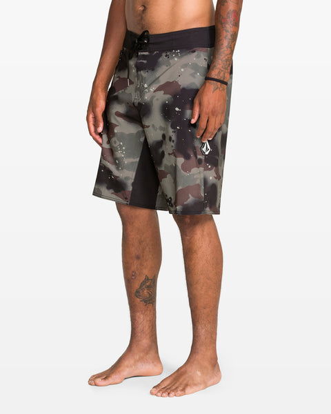 A person stands barefoot in Volcom Men's Fomenter Mod black and gray camo board shorts with a drawstring waist, made from REPREVE recycled fibers. Their left arm and leg have tattoos. The background is plain white.