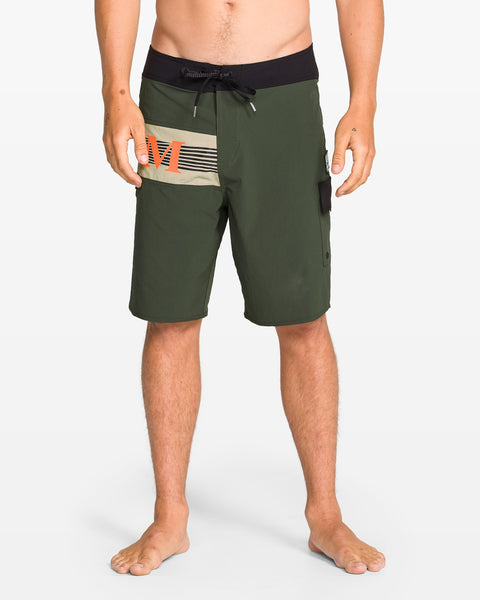 A person stands barefoot in Volcom Men's Liberator Mod olive green 20" board shorts with 4-way stretch nylon, black waistband with Cinch Fly Technology, drawstring, side pocket, and beige/orange stripes on the left thigh.