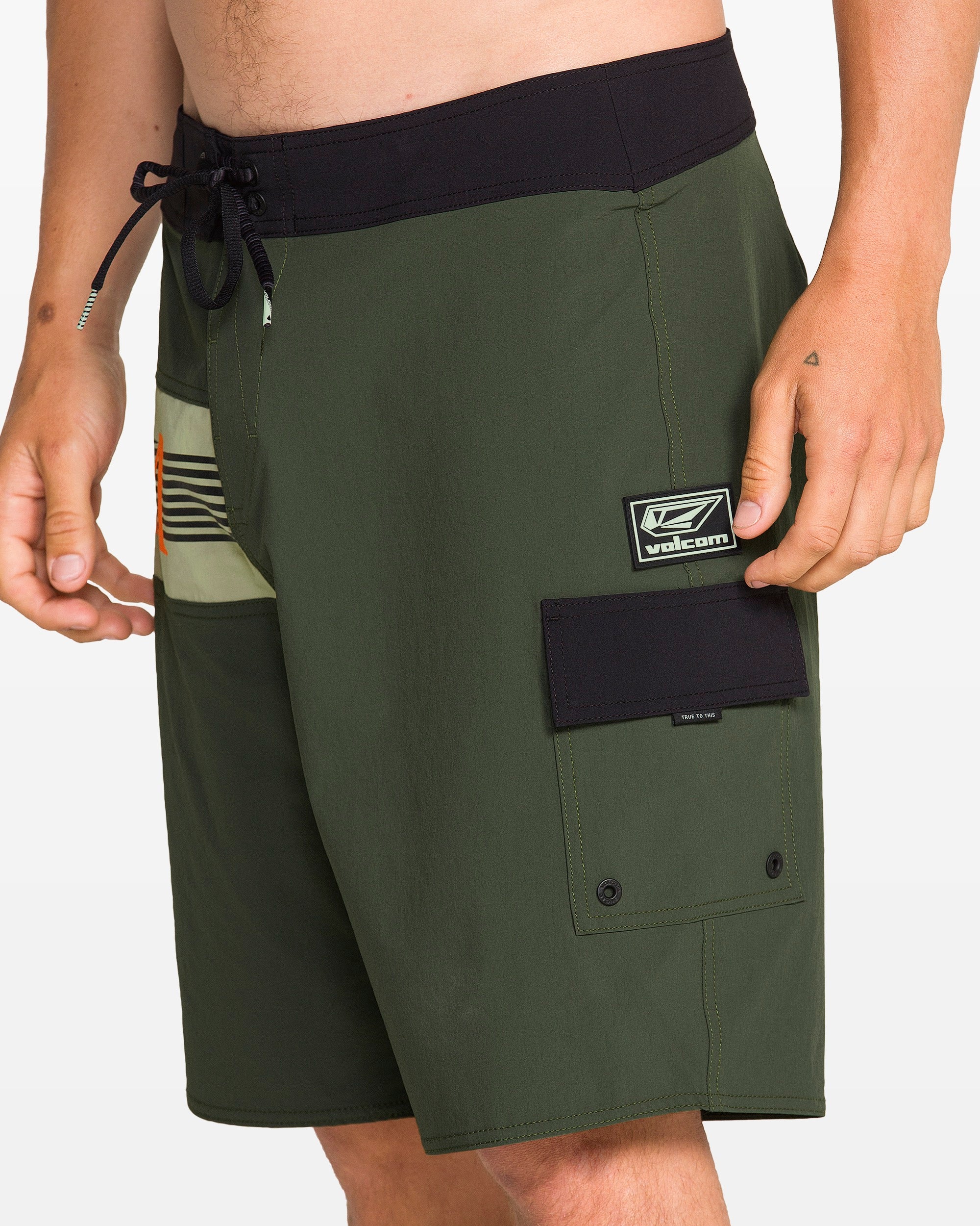 A person wears Volcom Men's Liberator Mod board shorts in dark green with a black waistband and pocket, logo patch, drawstring waist, and 4-way stretch nylon. Only the torso and upper legs are shown.