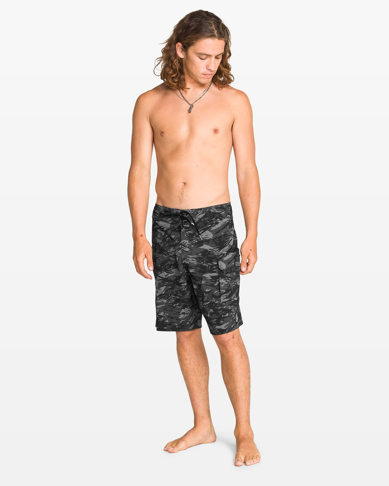 A barefoot man with long hair stands against a white background, wearing Volcom Men's 4th Of July Preemo Mod boardshorts featuring Cinch Fly Technology and a necklace. He is shirtless and looking down to his left.