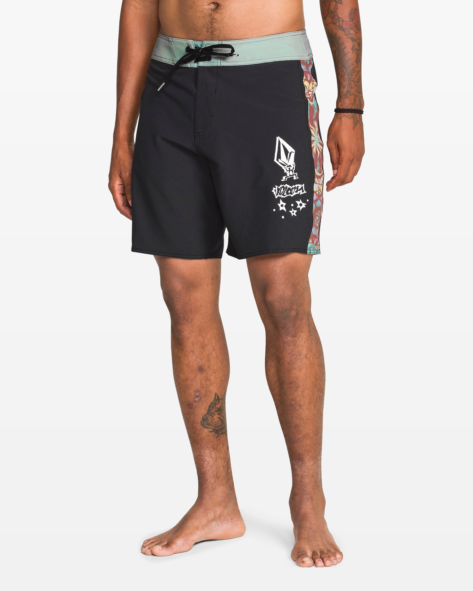 A person stands barefoot wearing Volcom Men's Star Shields Featured Artist Mod boardshorts with a green waistband, multicolored side design, left thigh graphics, and Cinch Fly Technology. Their right arm and left leg are tattooed.