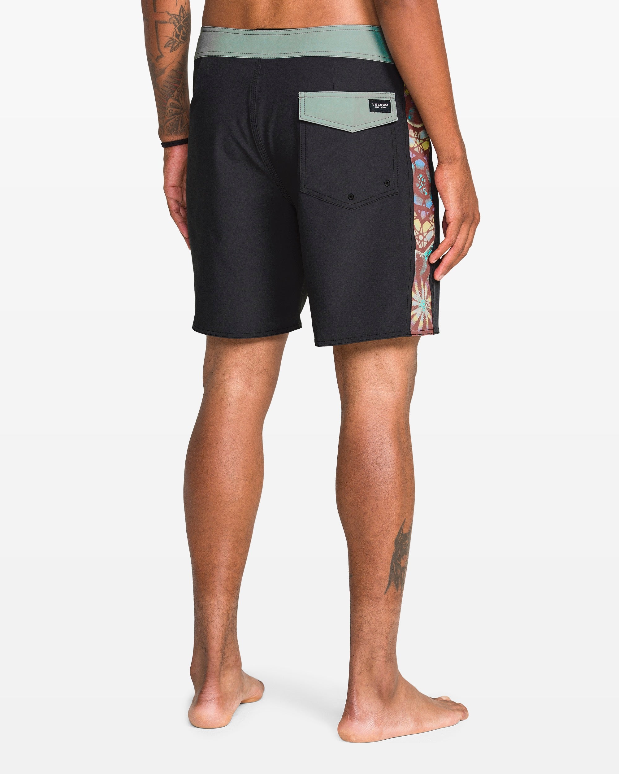 A person with a leg tattoo and medium skin tone stands barefoot, facing away, wearing Volcom Men's Star Shields Featured Artist Mod boardshorts with a green waistband, back flap pocket, and colorful patterned side panel.