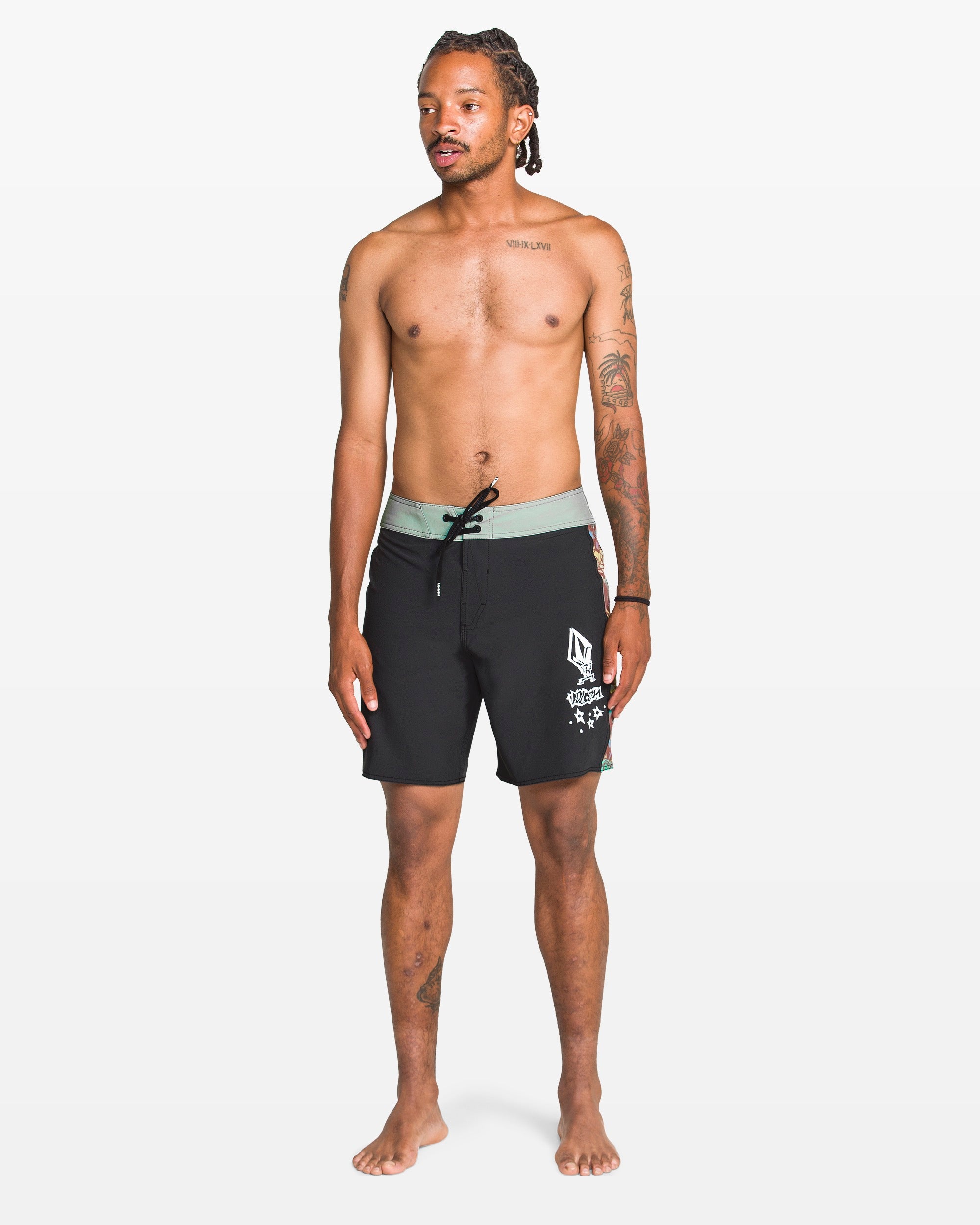 A barefoot, tattooed man stands against a white background wearing Volcom Men's Star Shields Featured Artist Mod boardshorts with a green waistband. He is shirtless, looking to the side, and captures the unique vibe of the Featured Artist Series.