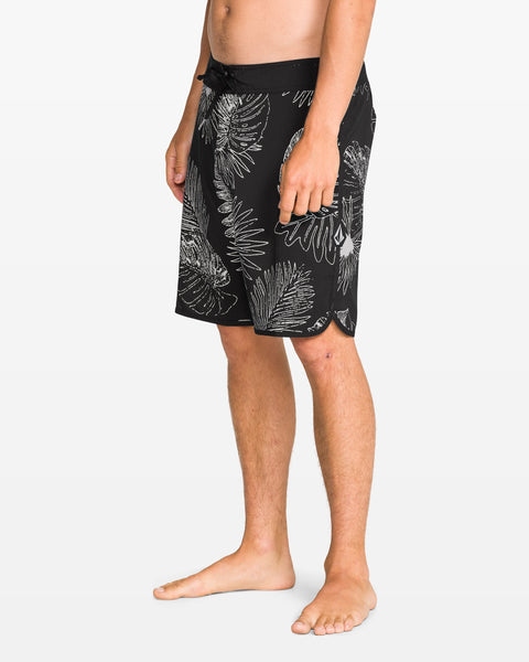 A person stands sideways, barefoot against a white background, wearing Volcom Men's Infuse Scallop Mod 19" boardshorts featuring a black and white tropical leaf print, Cinch Fly Technology, scalloped hem, and a drawstring waist.