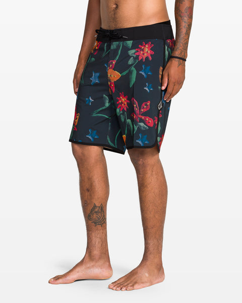 A man stands barefoot on a white background wearing Volcom’s Men's Infuse Scallop Mod boardshorts with a colorful floral pattern; his right arm and left leg are tattooed, while Cinch Fly Technology keeps them secure.