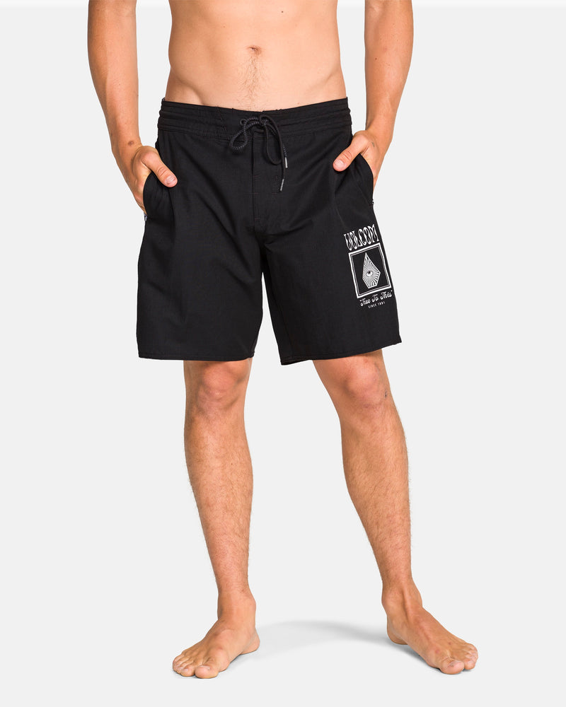 A person wears black Volcom Men's Yewster Loose Stoney boardshorts featuring a white graphic on the left leg and pockets, made with REPREVE recycled fibers, standing barefoot against a plain background.