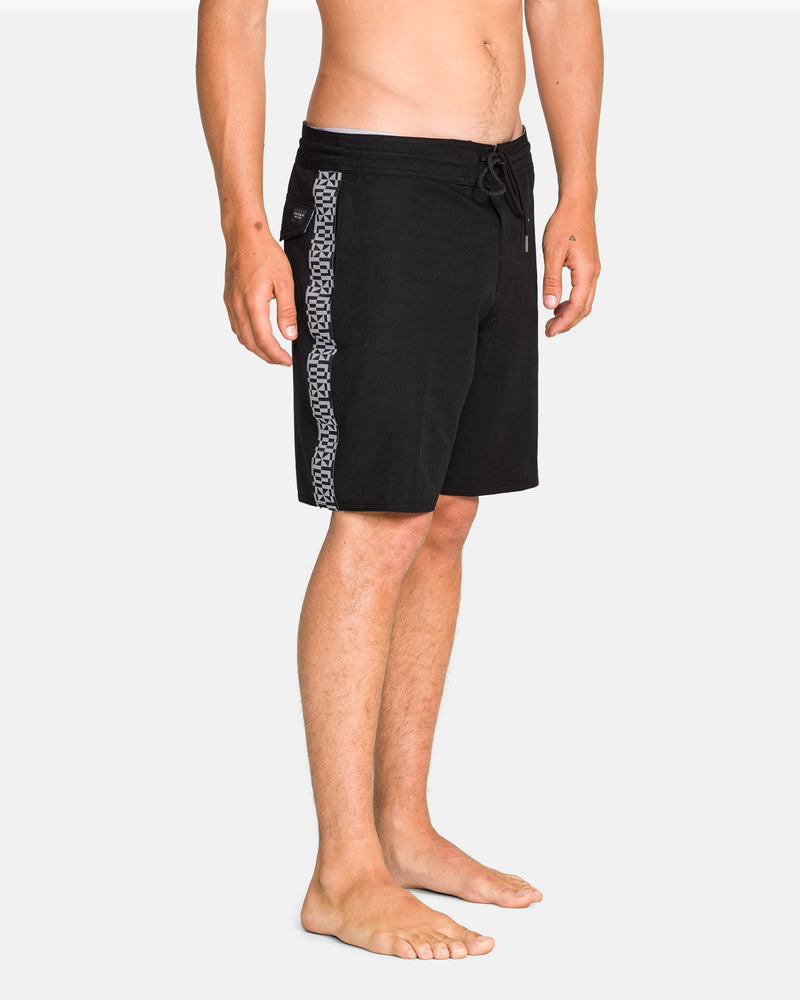 A man stands barefoot against a white background, wearing Volcom Men's Yewster Loose Stoney black knee-length boardshorts featuring a geometric patterned stripe down the sides and made with REPREVE recycled fibers.