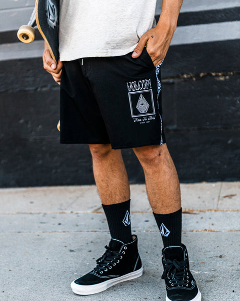 A person wearing Volcom Men's Yewster Loose Stoney black boardshorts, black socks, and black sneakers stands on a sidewalk holding a skateboard. Only the lower half of their body is visible.