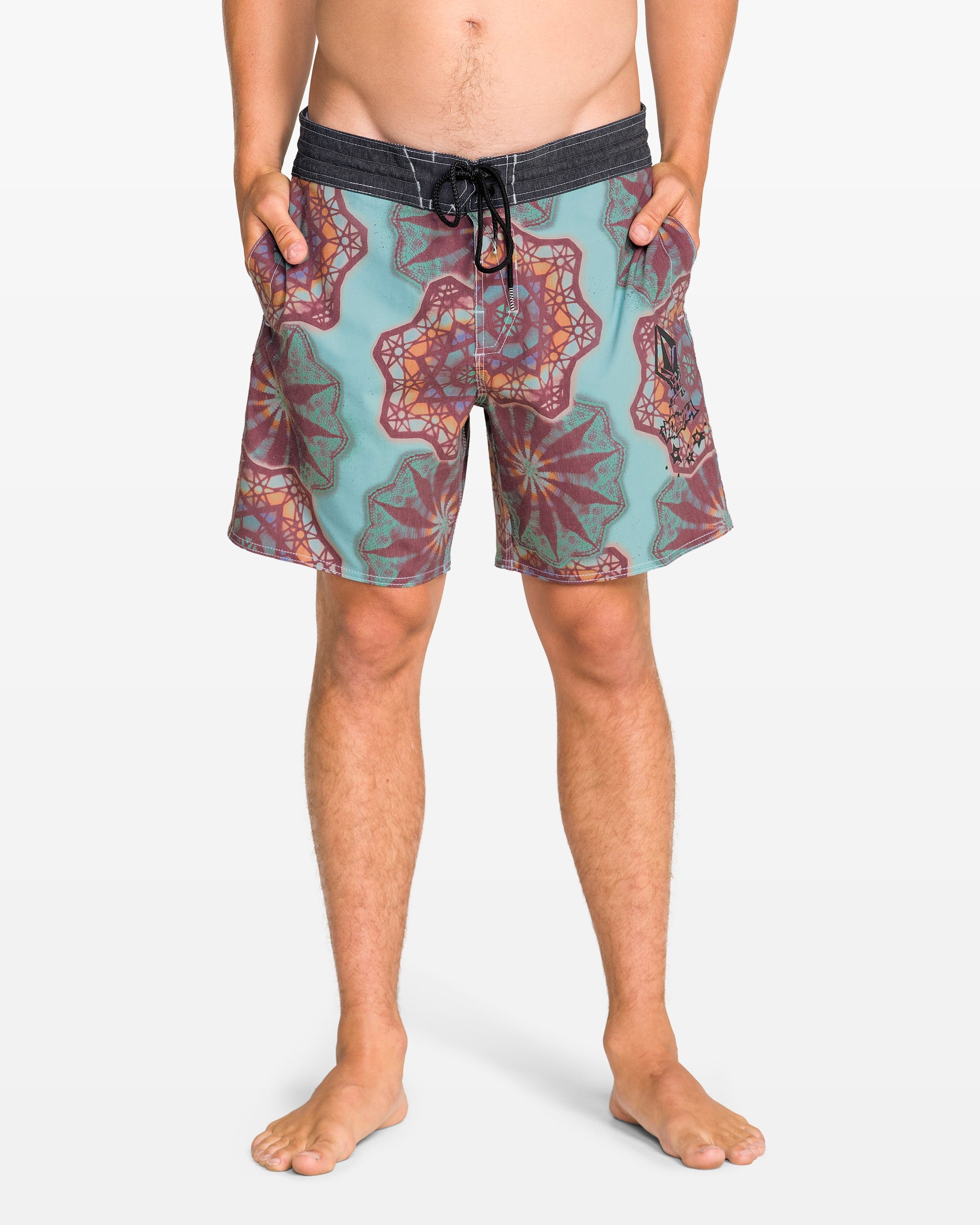 A man stands barefoot with hands in pockets, wearing Volcom Men's Star Shields Featured Artist Pcs Stoney trunks in teal and maroon patterns with a black waistband. This standout style pops against the plain white background.