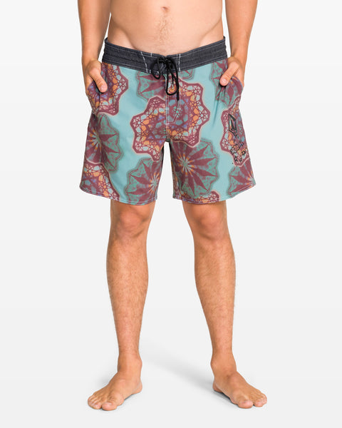 A man stands barefoot with hands in pockets, wearing Volcom Men's Star Shields Featured Artist Pcs Stoney trunks in teal and maroon patterns with a black waistband. This standout style pops against the plain white background.