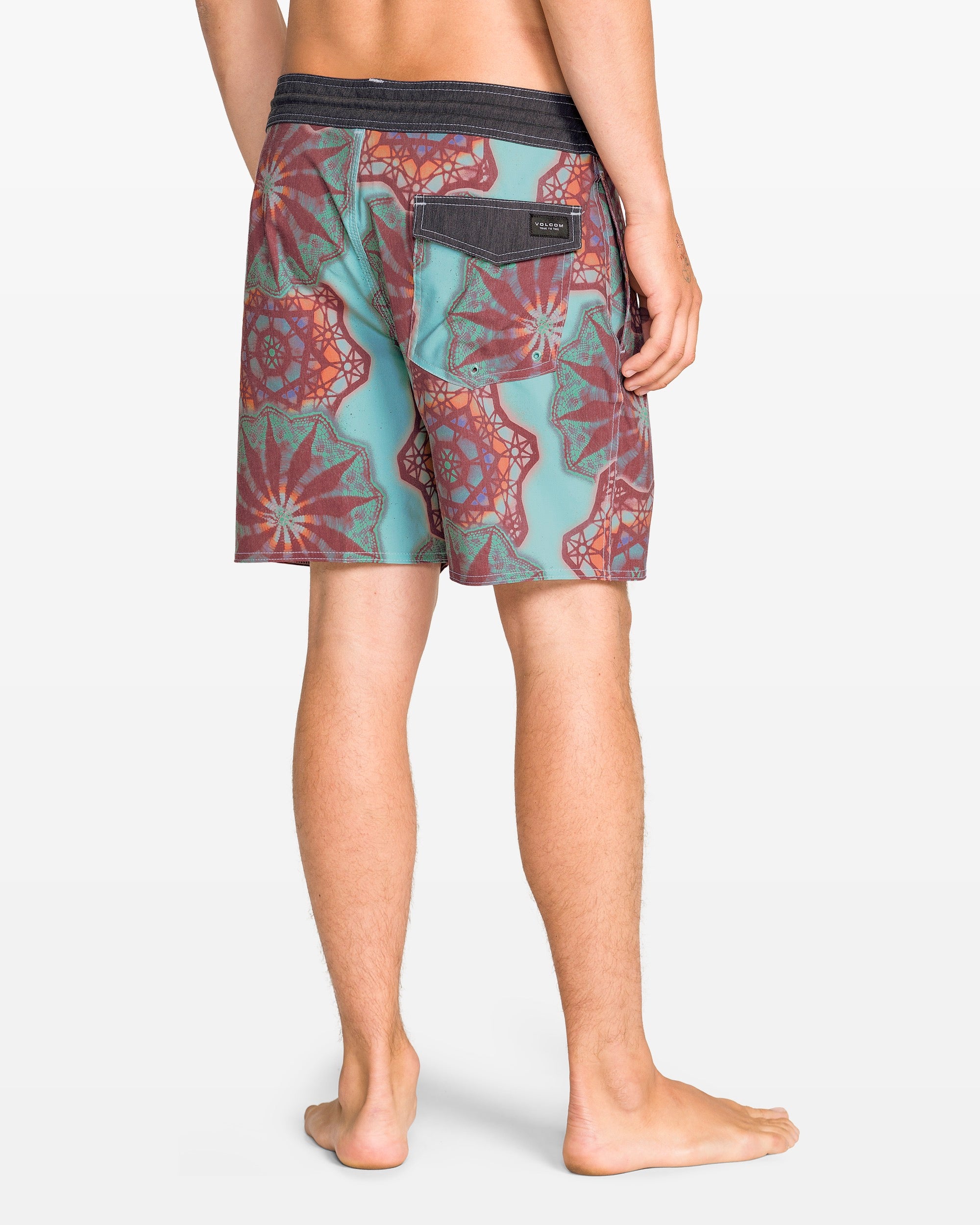 A person stands barefoot, shown from behind, wearing Volcom Men's Star Shields Featured Artist Pcs Stoney trunks with a black waistband and back flap pocket. The aquatic-ready shorts feature teal and red patterns against a plain white background.