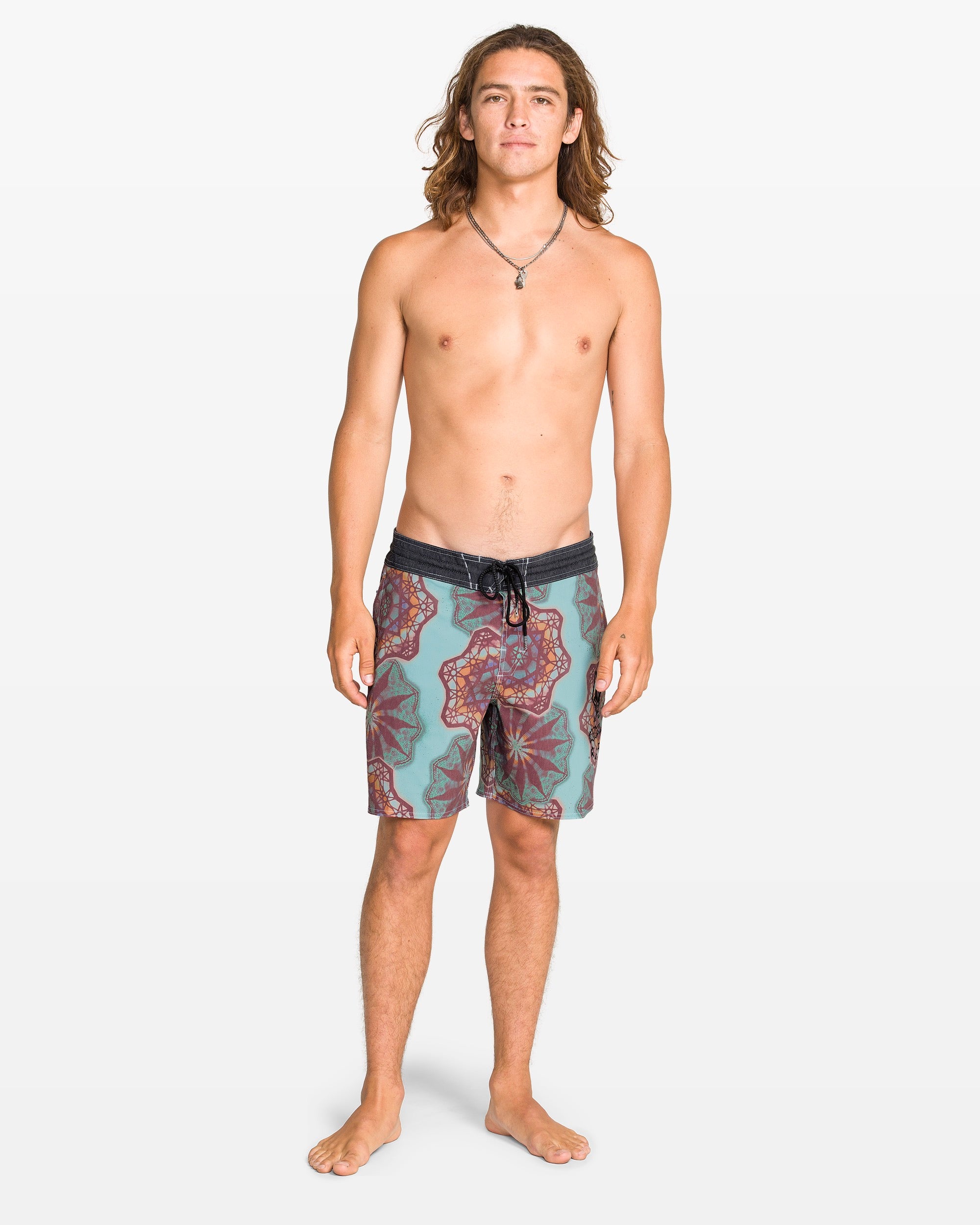 A young man with long brown hair stands barefoot, wearing Volcom Men's Star Shields Featured Artist Pcs Stoney trunks and a necklace, facing forward against a plain white background—an aquatic-ready look.