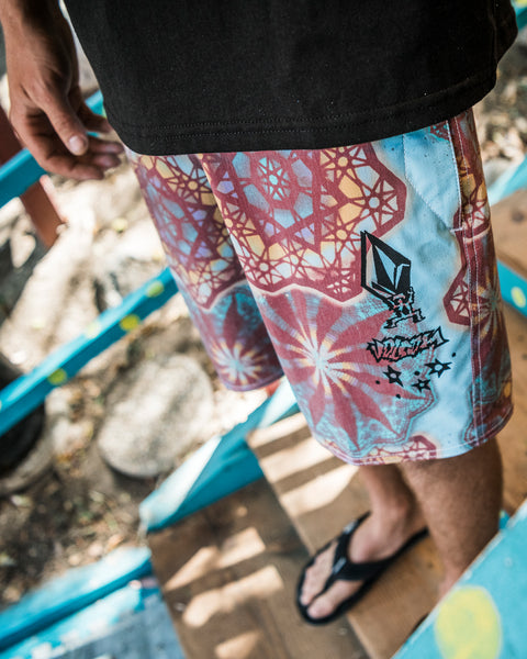 A person wearing Volcom Men's Star Shields Featured Artist Pcs Stoney trunks with red, yellow, and blue star designs, a black shirt, and flip-flops stands on painted wooden steps outdoors.