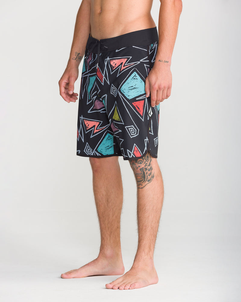 A man stands barefoot wearing Volcom Men’s Infuse Scallop Mod 19 boardshorts in black with colorful geometric designs. The shorts feature 4-way stretch for maximum comfort and flexibility. He is shirtless, revealing tattoos on his left forearm and right upper leg, against a plain light-colored background.