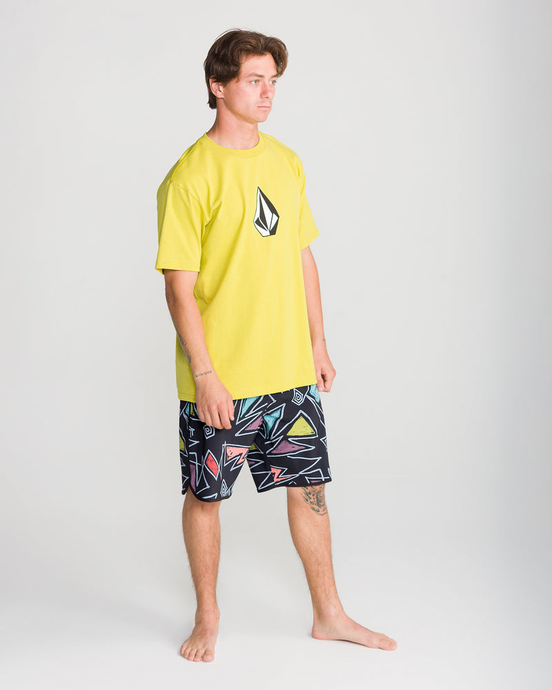 A man stands barefoot on a plain white background, wearing Volcom’s Men’s Infuse Scallop Mod 19 boardshorts featuring 4-way stretch and a yellow T-shirt with a black logo, looking slightly to the side.