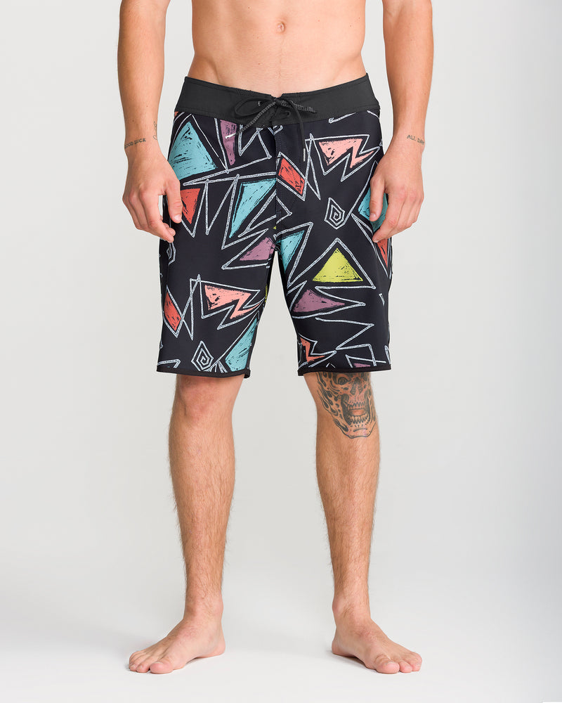 A barefoot man models the Volcom Men’s Infuse Scallop Mod 19 boardshorts, featuring a bold geometric pattern with triangles and zigzags in pink, teal, yellow, and orange. Designed with advanced Cinch Fly Technology and 4-way stretch fabric for maximum comfort and flexibility, these boardshorts showcase his bare torso as well as tattoos on his arms and left leg.