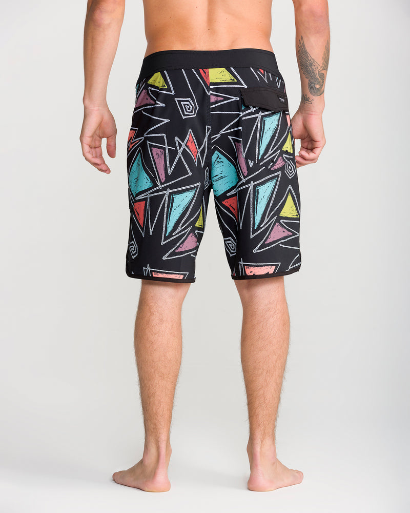 A man stands barefoot facing away, wearing Volcom Men’s Infuse Scallop Mod 19 boardshorts with a black waistband, colorful geometric patterns, and 4-way stretch for maximum comfort.