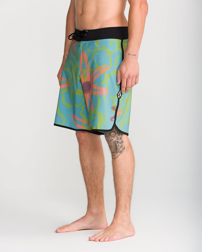 A person is wearing Volcom Men’s Infuse Scallop Mod 19 boardshorts featuring a floral pattern, 4-way stretch fabric, and a black waistband, standing barefoot on a white background. Only the torso and legs are visible, with a tattoo visible on one arm. The shorts have a 19-inch outseam and incorporate Cinch Fly Technology for enhanced comfort and flexibility.