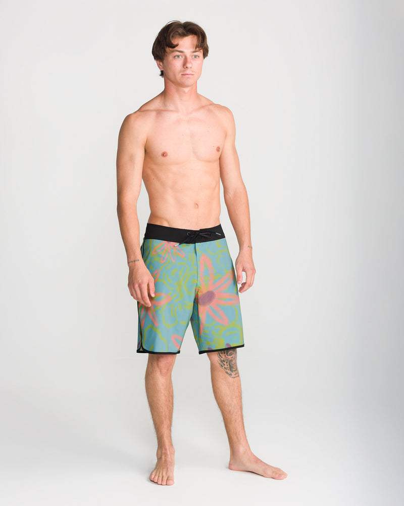 A young man with short brown hair and a tattoo on his left shin stands barefoot against a plain background, wearing only the Volcom Men’s Infuse Scallop Mod 19 boardshorts. These colorful shorts feature abstract patterns, a comfortable 19-inch outseam, and 4-way stretch fabric, while Cinch Fly Technology ensures they stay securely in place.