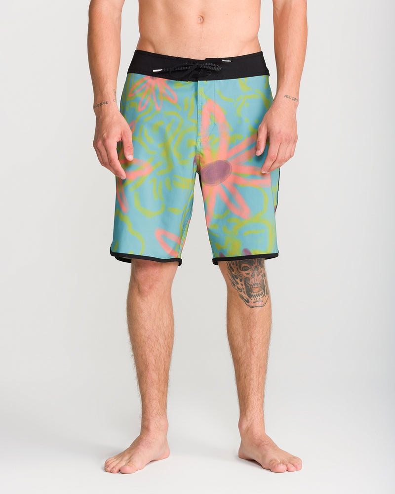 A shirtless person stands barefoot, facing forward and showcasing the Volcom Men’s Infuse Scallop Mod 19 boardshorts, featuring a black waistband, Cinch Fly Technology, and a drawstring for a secure fit. The colorful 4-way stretch shorts display pink floral and green patterns. A tattoo is visible on their right knee.