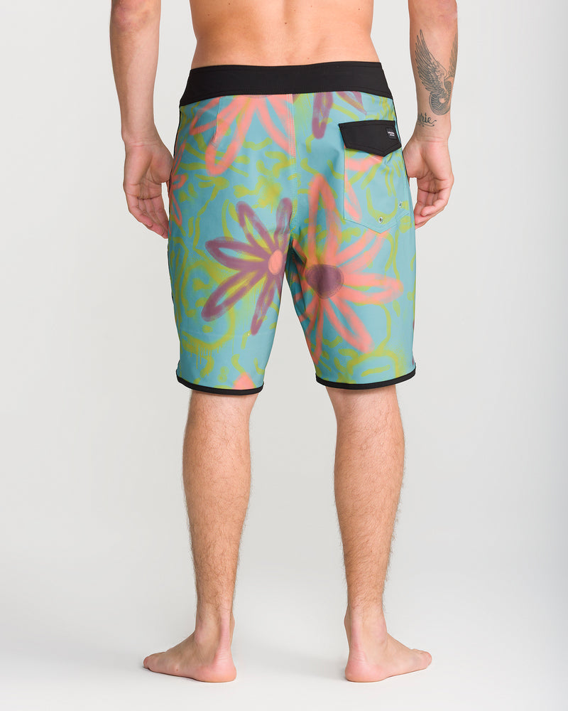 A barefoot person stands facing away, wearing Volcom Men’s Infuse Scallop Mod 19 boardshorts—colorful, knee-length with a floral print, featuring a black waistband with Cinch Fly Technology and a back pocket. A tattoo is visible on the right forearm. Set against a plain, light-colored background, the modern design and 19-inch outseam of these Volcom boardshorts are highlighted.