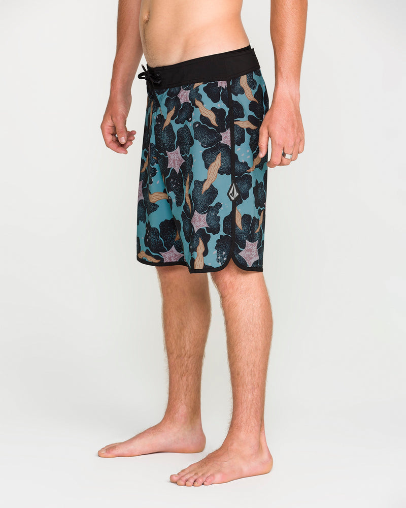 A person stands barefoot on a white background, wearing Volcom Men's Infuse Scallop Mod board shorts featuring a teal and black pattern, drawstring waist, and 4-way stretch for maximum mobility. Only the lower half of the body is visible.