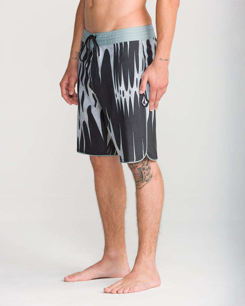 A man stands barefoot, wearing Volcom Men’s Barnacle Stoney 19 boardshorts with a black and gray pattern, drawstring waist, and stylish scallop cut. His left arm and leg feature visible tattoos. The background is plain and light-colored.