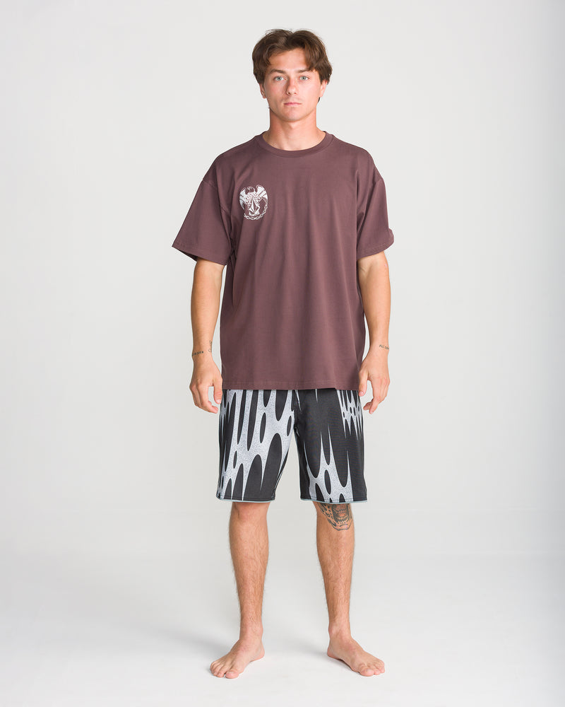 A person stands barefoot on a white background, wearing a loose brown T-shirt with a small white graphic on the chest and Volcom Men’s Barnacle Stoney 19 boardshorts, which feature a bold white pattern and a modern scallop cut.