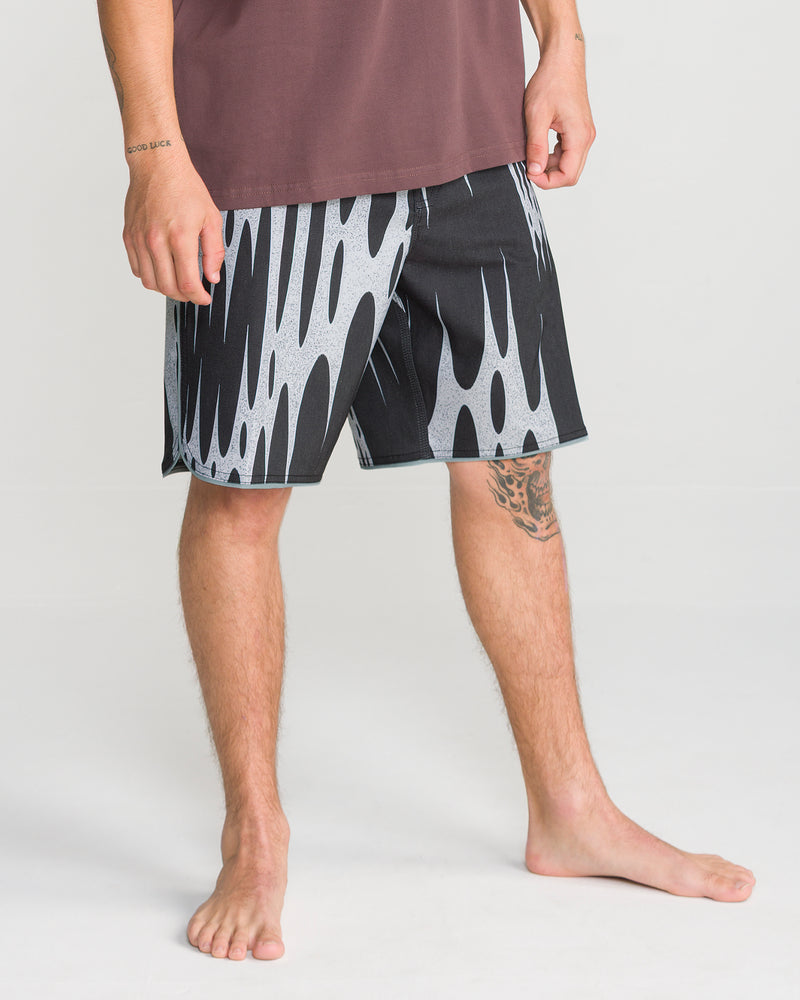 A person stands barefoot on a white floor, wearing the Volcom Men’s Barnacle Stoney 19 boardshorts featuring a black-and-white flame pattern and scallop cut; only the lower half of their body is visible.