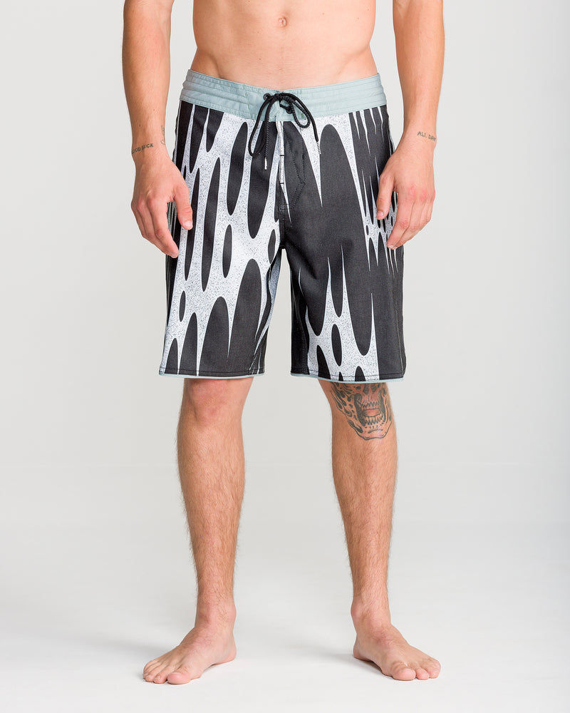 A barefoot person wearing Volcom Men’s Barnacle Stoney 19 boardshorts, featuring a black and white pattern with a scallop cut and light waistband, stands against a plain light background. Tattoos are visible on the right thigh and both forearms.