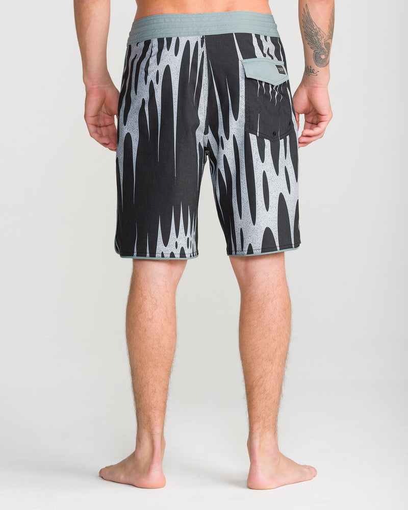 A person stands barefoot facing away, wearing Volcom Men’s Barnacle Stoney 19 black and gray patterned boardshorts with a scallop cut, featuring a small back pocket and distinct waistband detail.