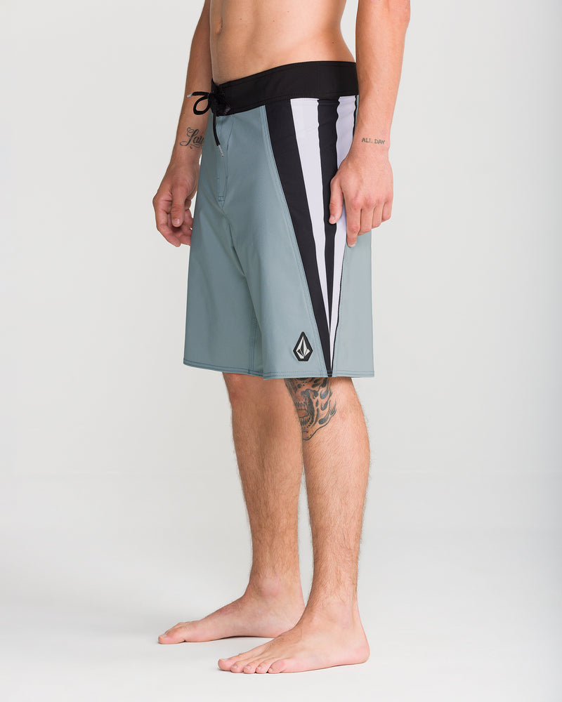 A man stands barefoot wearing Volcom’s Men’s Facet Mod 20 boardshorts in light blue, featuring black and white side panels, a black waistband, and 4-way stretch fabric for enhanced comfort. Visible tattoos adorn his left forearm and right leg. The background is plain and light-colored.