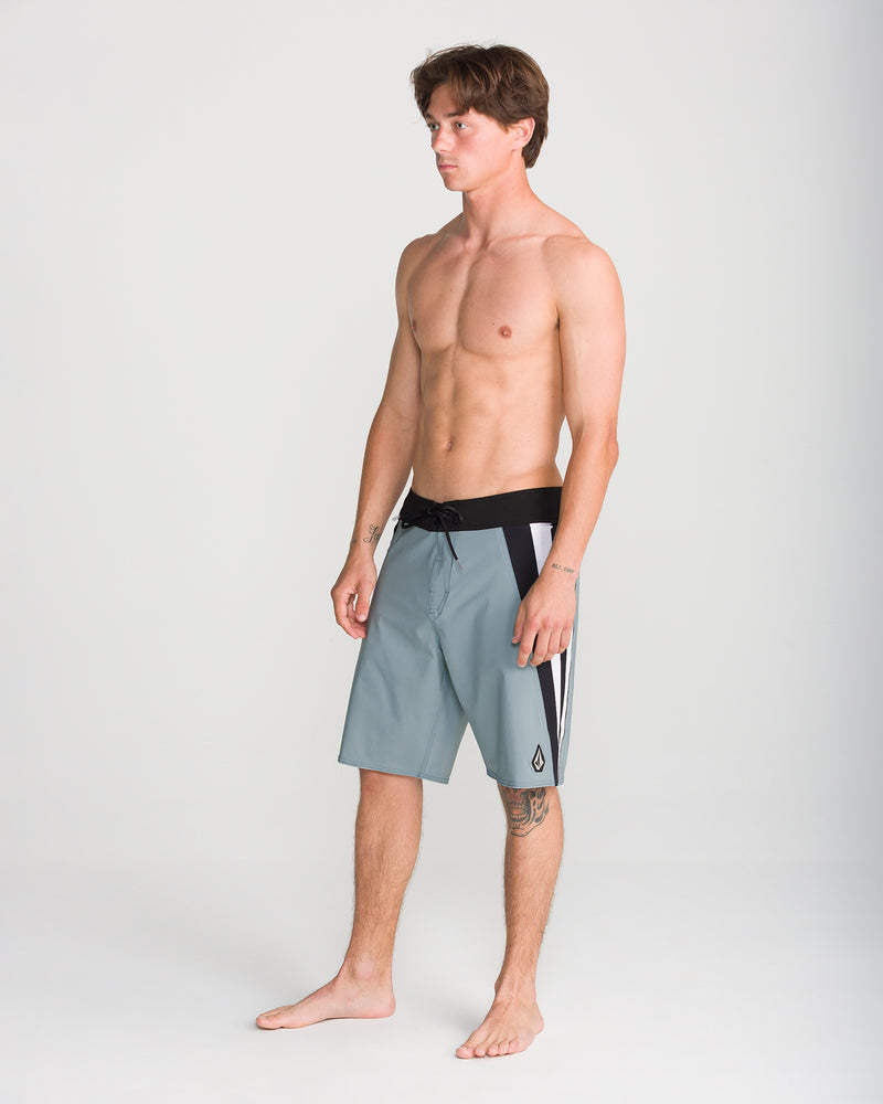A young man stands barefoot on a plain backdrop, wearing Volcom Men’s Facet Mod 20 boardshorts in gray, featuring black and white side panels. He is shirtless, facing slightly to the left, with short brown hair. The boardshorts are made with 4-way stretch fabric for maximum comfort and flexibility.