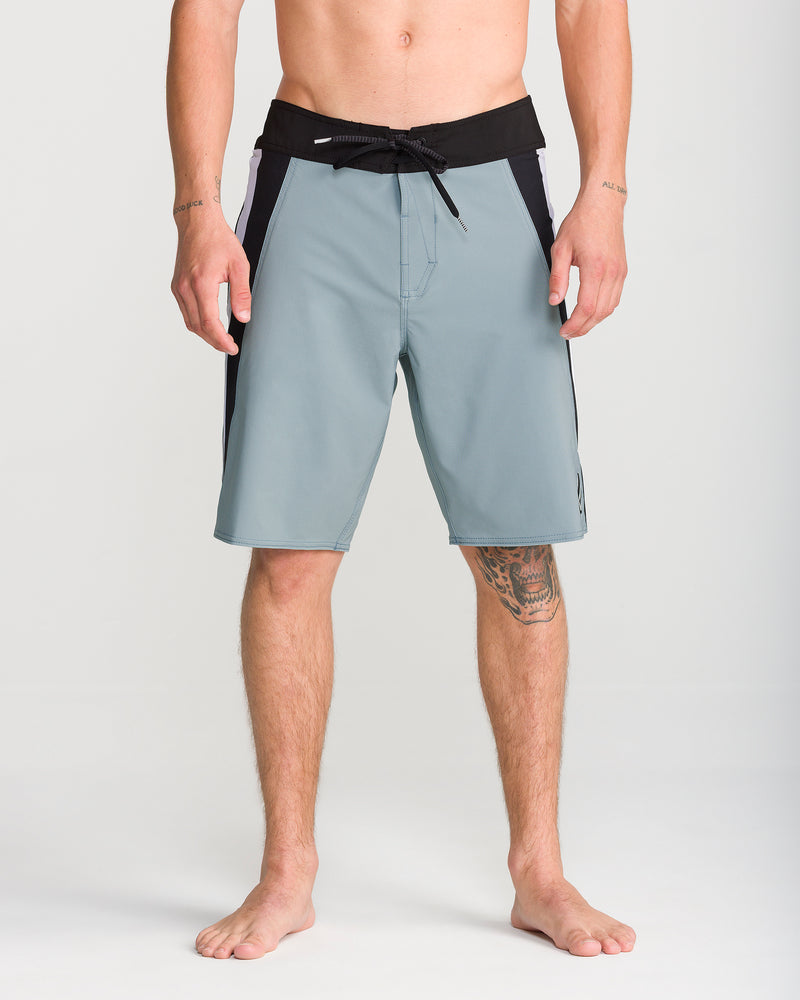 A man stands barefoot against a plain light background, wearing Volcom Men’s Facet Mod 20 boardshorts in light blue with a 4-way stretch, featuring a black waistband and black side panels; his hands are relaxed by his sides, and a tattoo is visible on his right thigh.