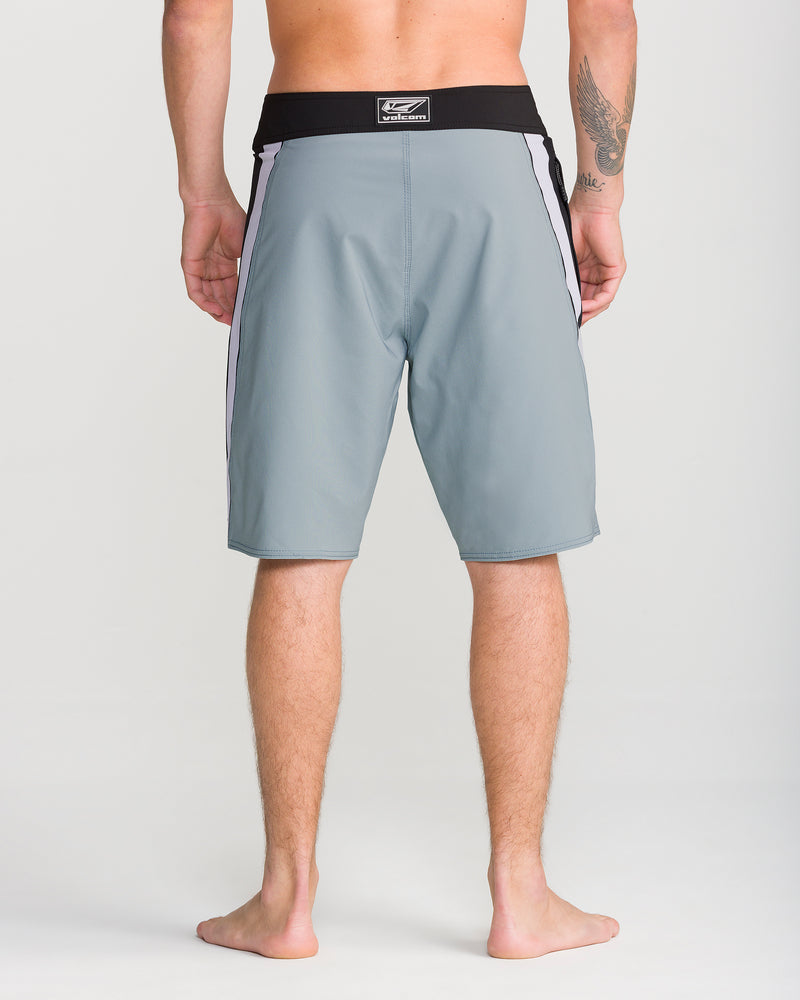 A barefoot man stands facing away, wearing Volcom Men’s Facet Mod 20 boardshorts in light gray with a black waistband and side stripes. He has a visible tattoo on his left upper arm.