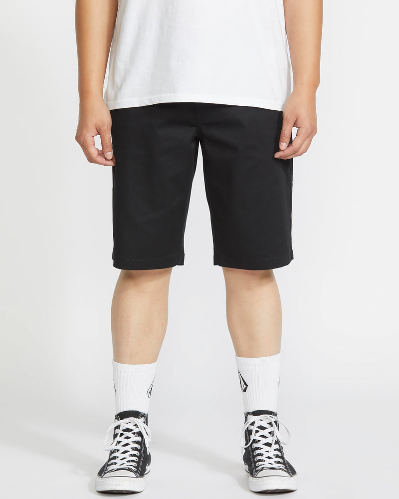 A person stands against a white background, wearing a Volcom white t-shirt, Volcom Men's Frickin Modern Stretch Shorts with 5-pocket styling, white crew socks, and black-and-white sneakers. The image is cropped at the shoulders.
