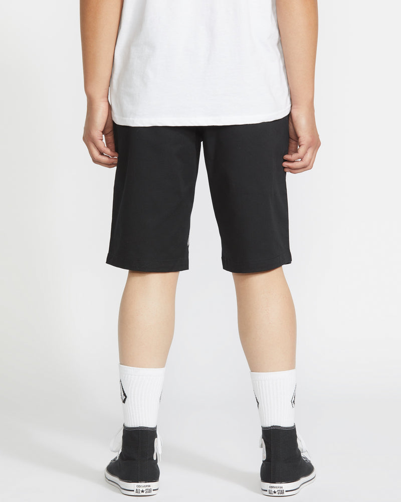 A person is facing away from the camera, wearing a white T-shirt, Volcom Men's Frickin Modern Stretch Shorts with 5-pocket styling, white socks, and black sneakers against a plain white background.