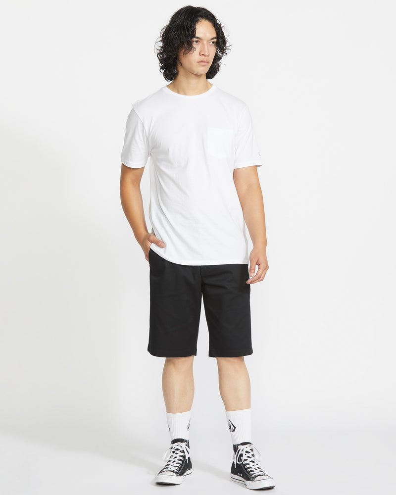 A person with curly hair wears a white T-shirt, Volcom Men’s Frickin Modern Stretch Shorts in black with 5-pocket styling, white socks, and black high-top sneakers, standing against a plain white background.