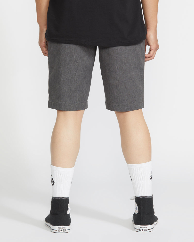 A person faces away from the camera, wearing a black shirt, Volcom Men's Frickin Modern Stretch Shorts in gray, white socks with a small black logo, and black sneakers with white soles against a plain white background.