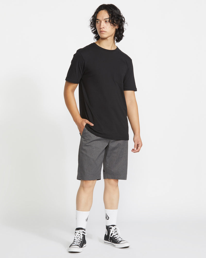 A person stands against a plain white background wearing a Volcom black t-shirt, Men's Frickin Modern Stretch Shorts by Volcom in breathable gray cotton, white socks with black logos, and black high-top sneakers. One hand rests in their pocket as they look sideways.