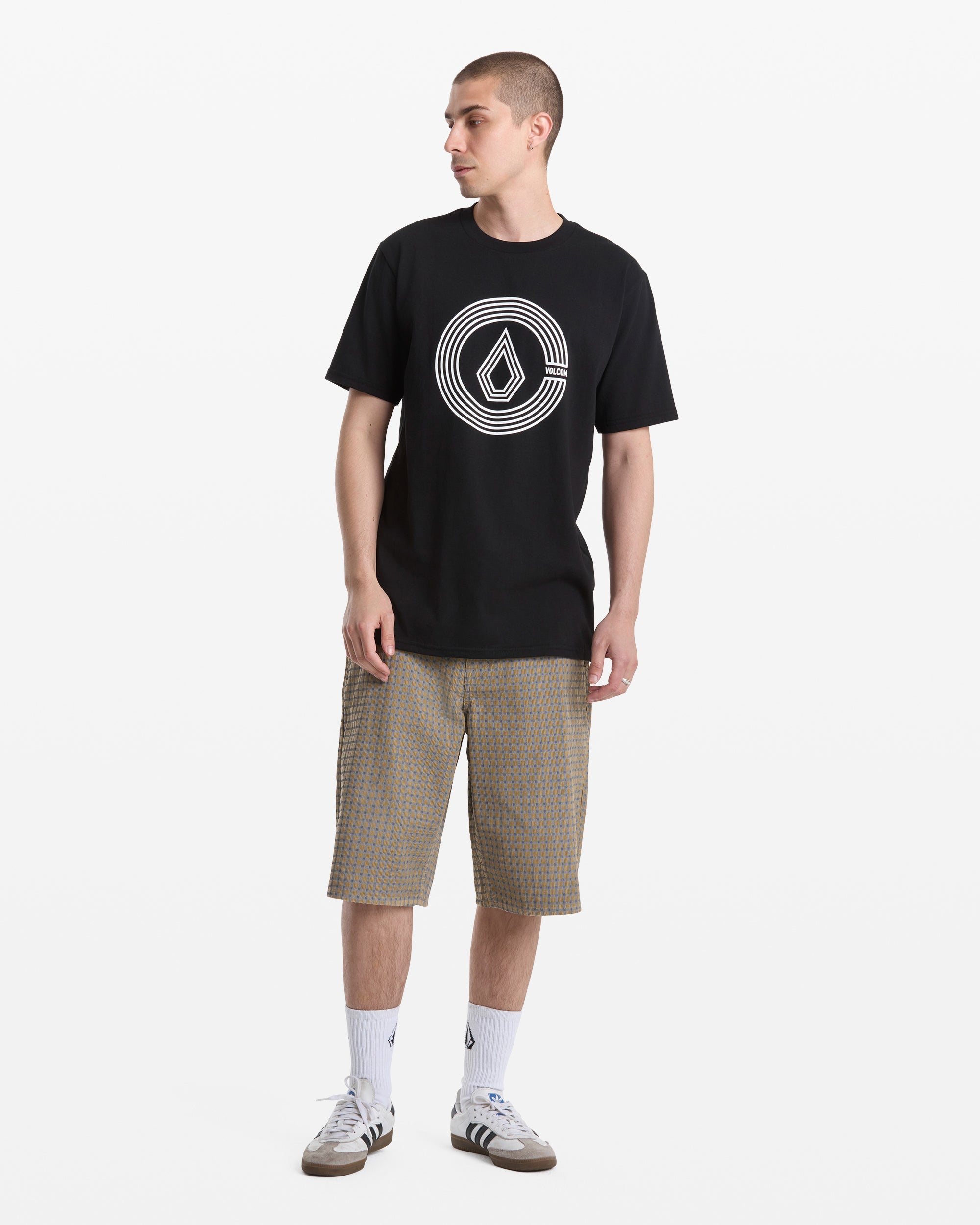 A man stands against a white background wearing Volcom Men's Chillow Shorts, a black graphic T-shirt, white crew socks, and gray Adidas sneakers. He looks to the side with his hands relaxed at his sides.