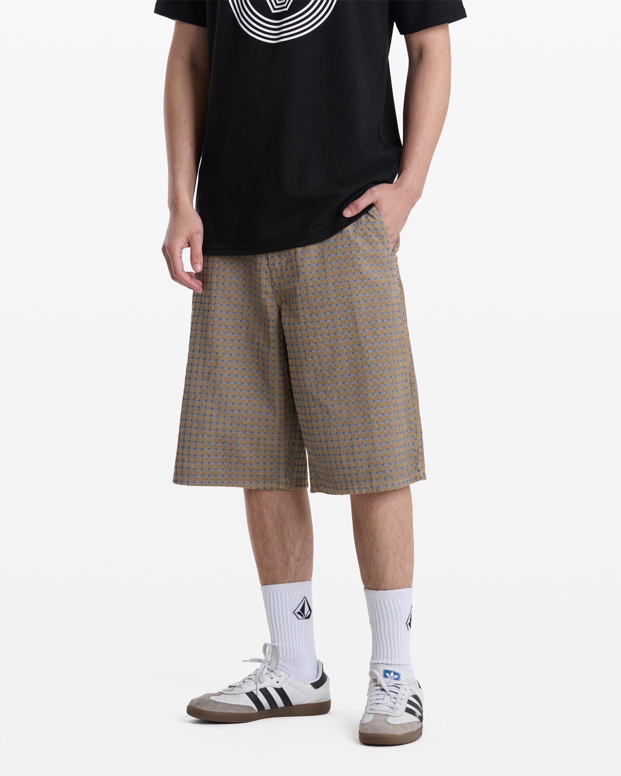A person wearing Volcom Men's Chillow Shorts, a black graphic tee, white crew socks with logos, and gray Adidas sneakers stands against a plain white background.