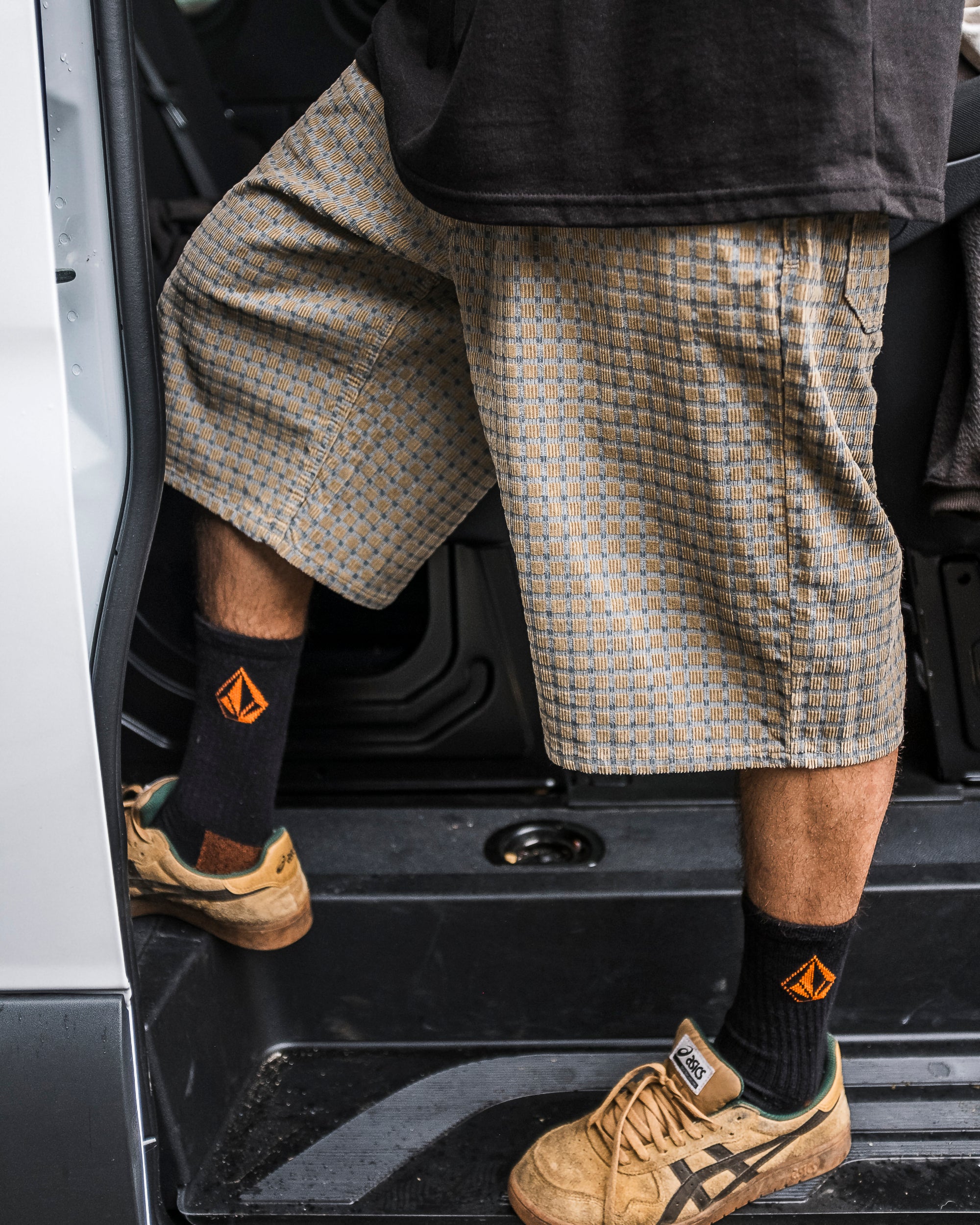 A person in Volcom Men's Chillow 24" Shorts with a loose fit, black socks with orange geometric patterns, and tan sneakers steps out of a car. Only the lower half is visible.