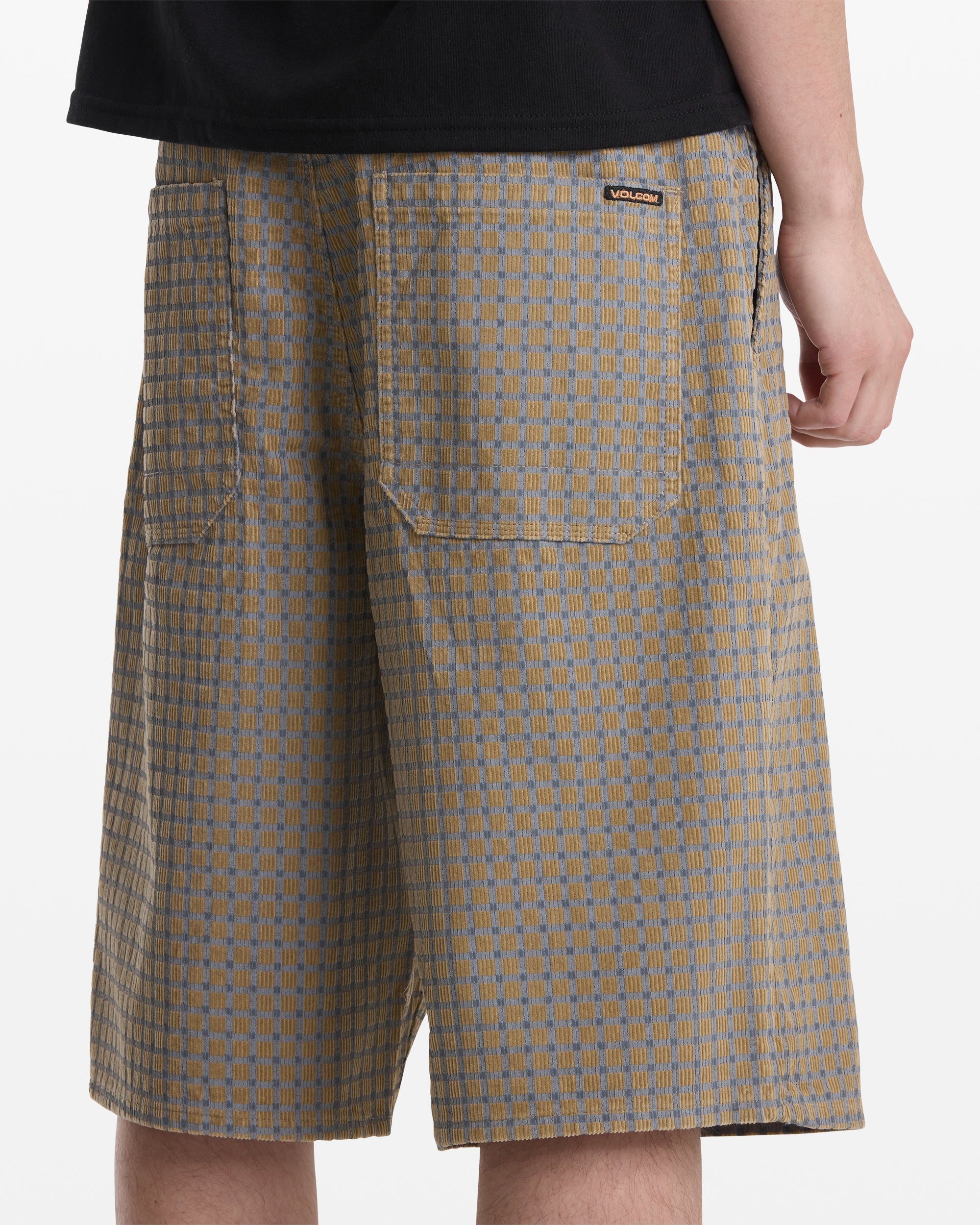 A man models the Volcom Men's Chillow Short—loose, knee-length shorts in beige and gray patterns with large back pockets and a small logo tag. Only his lower torso and legs are shown as he faces away.