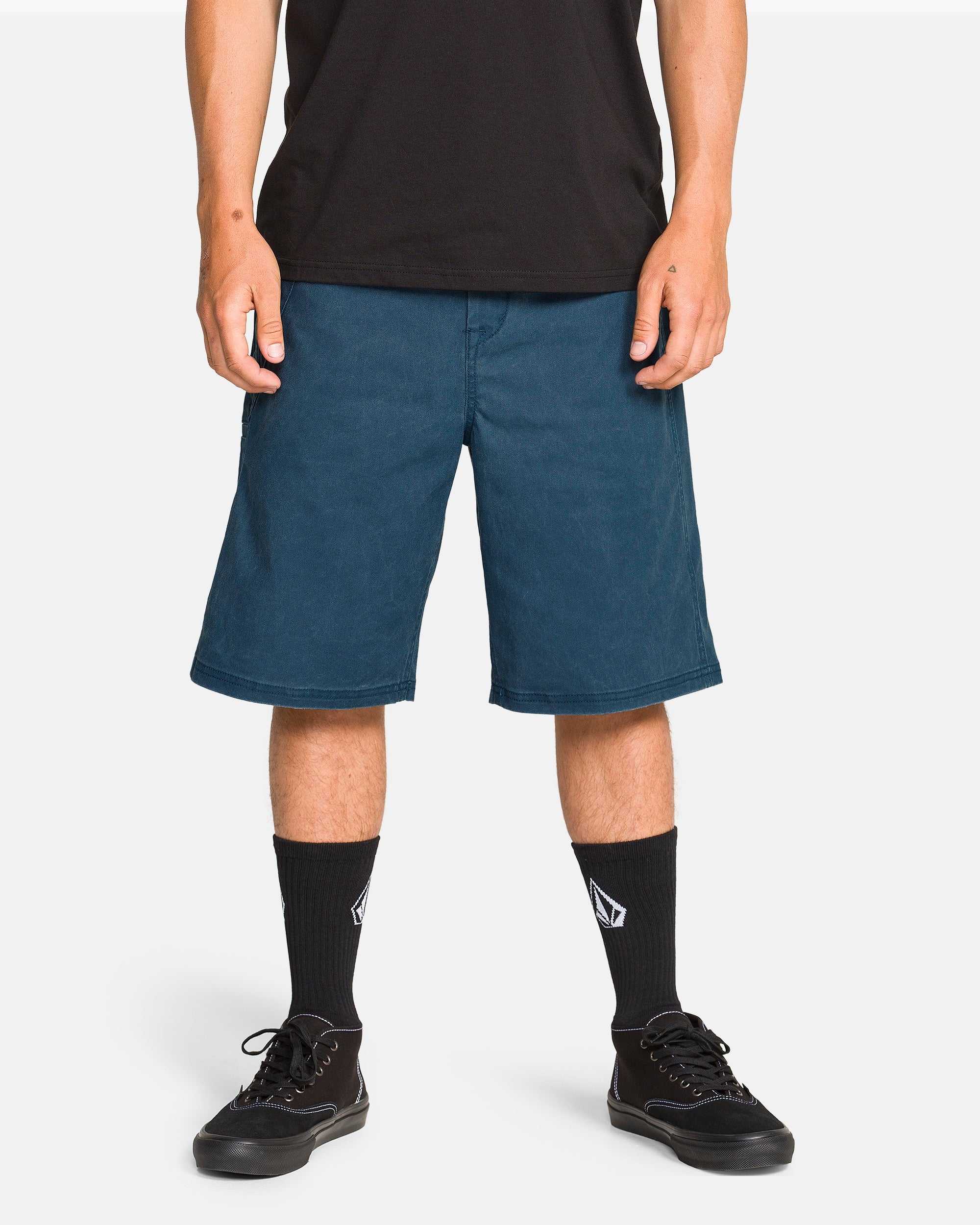 A person wears Volcom Men's Dialed Relaxed Work Shorts in blue with utility pockets, a black t-shirt, black sneakers, and black socks with a white logo, standing against a plain white background.